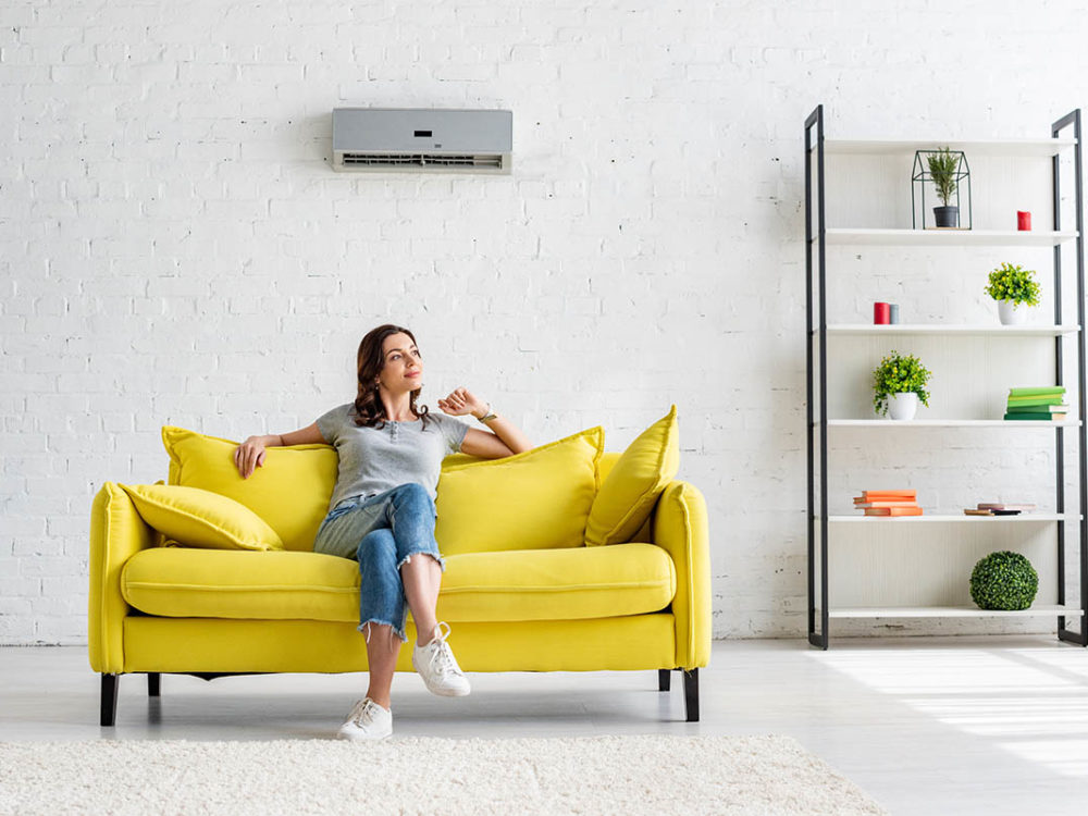 beautiful young woman sitting on yellow sofa under air conditioner in spacious apartment